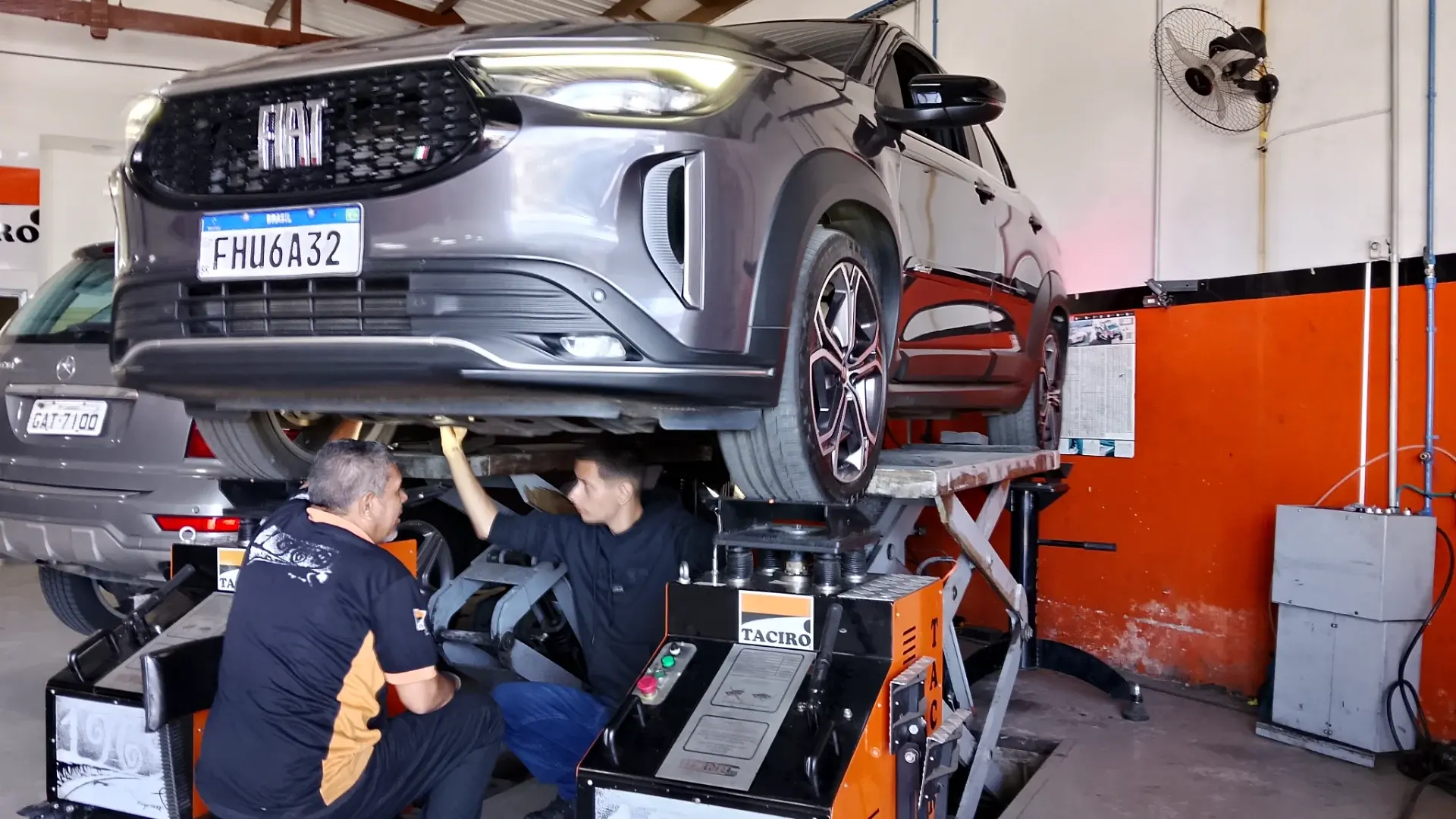 Dois mecânicos da Taciro embaixo de uma Fiat Fastback suspensa, operando a máquina de balanceamento no local