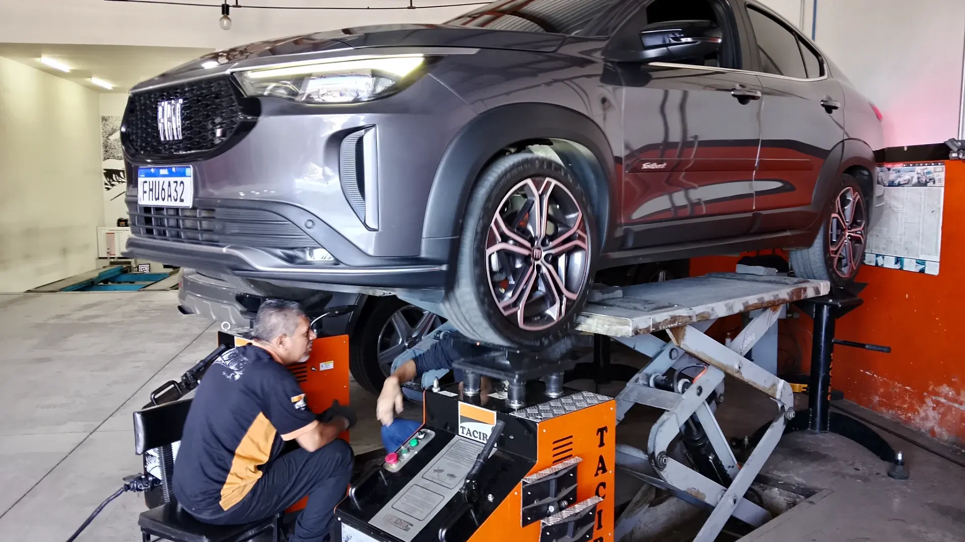 Fiat Fastback cinza suspensa no elevador tesoura da Taciro, com o mecânico sentado ao lado da máquina de balanceamento laranja da oficina